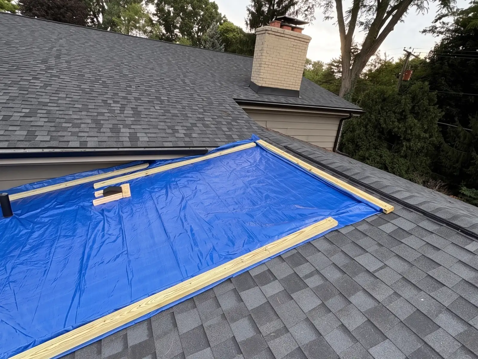 Blue emergency tarp nailed over a section of a shingle roof near a brick chimney