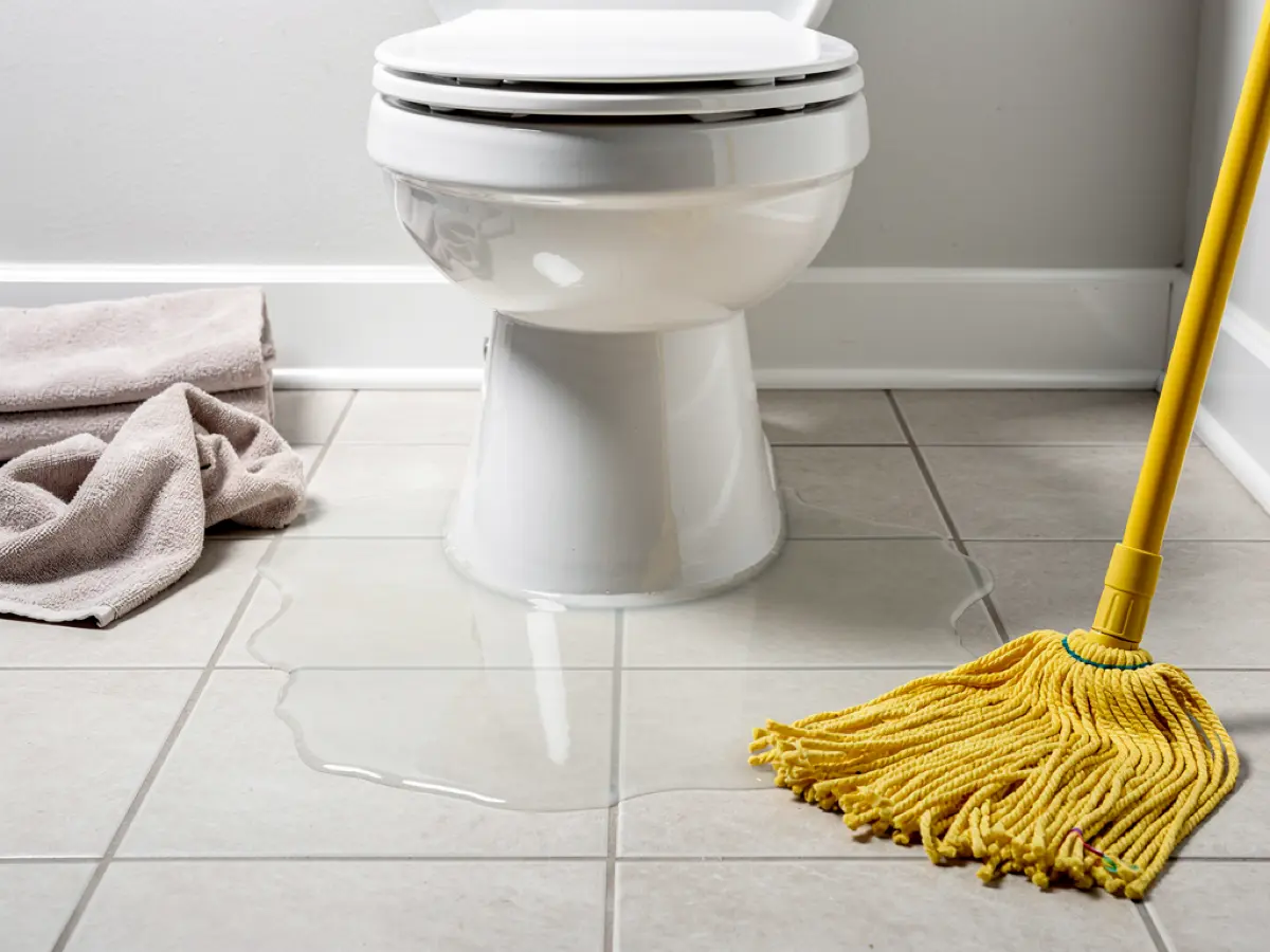 Bathroom tile floor with water at the base of a toilet and a mop and towels nearby