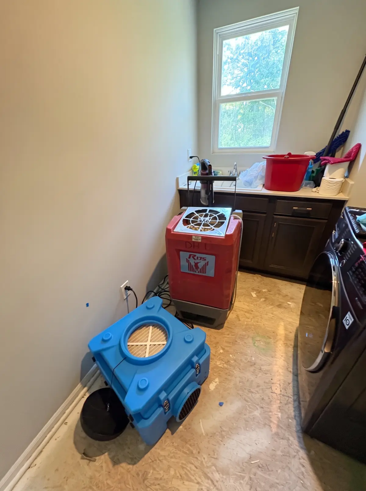 Laundry room with a dehumidifier and air mover running on an exposed subfloor beside a washer and dryer