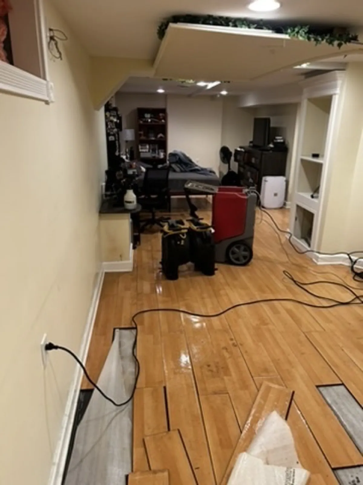 Basement ceiling opened near a doorway, with hardwood flooring pulled back and an air mover in place